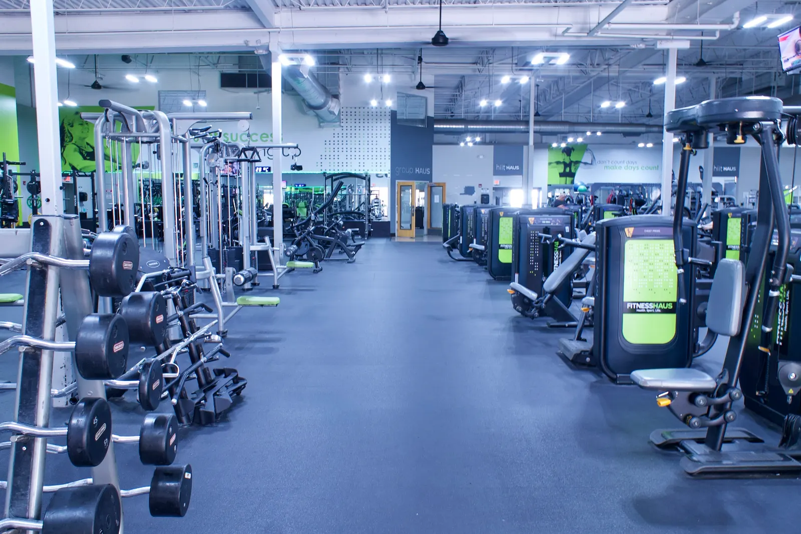 The machine floor looking down a long aisle, with dumbbells racked along one side and screens overhead.