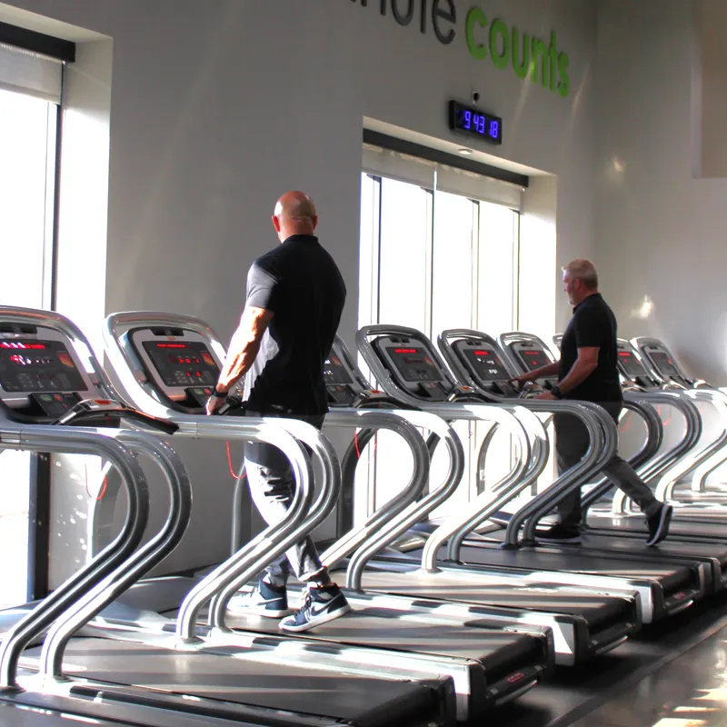 Two men walking on treadmills beside tall windows, seen from behind.