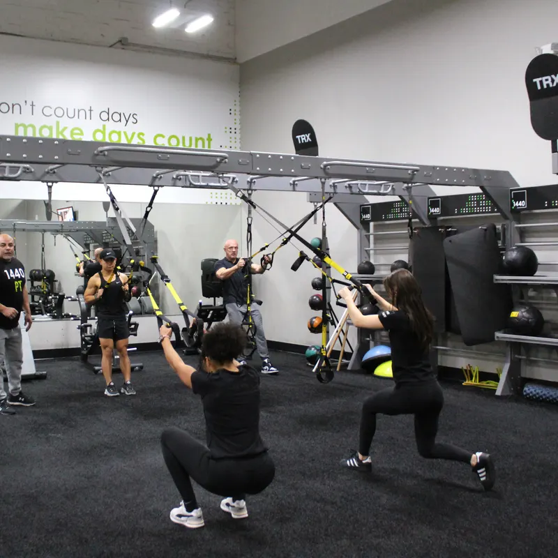 Two women training on suspension straps on the turf, one in a deep squat and one in a lunge.